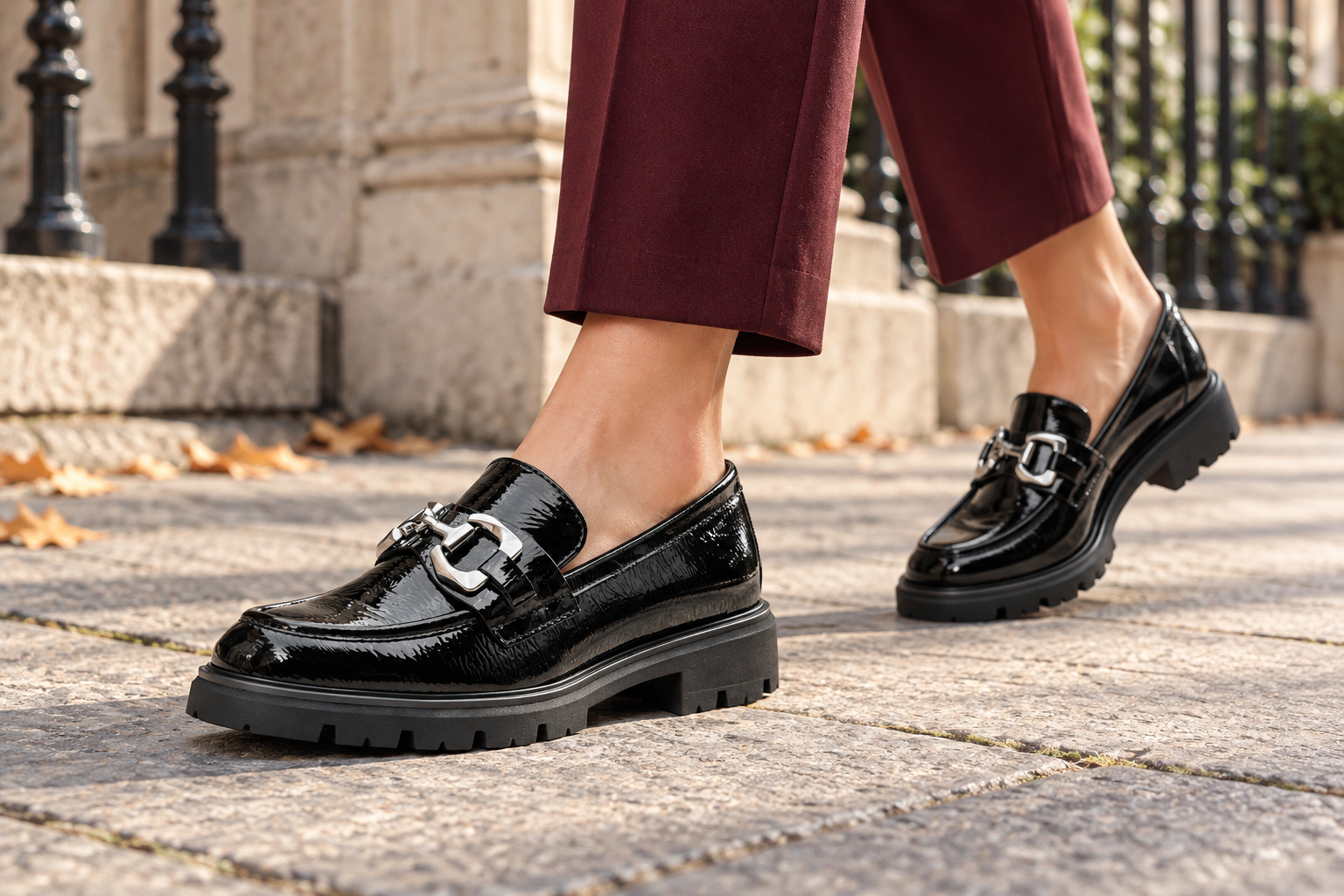 Close-up of black patent chunky loafers with silver trim, worn with burgundy trousers on a stone city pavement