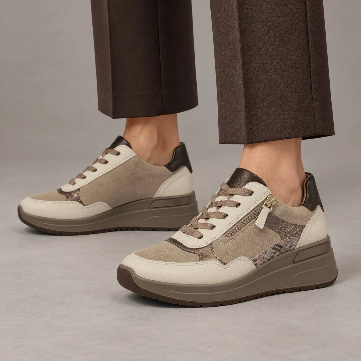 A pair of low-top platform sneakers in beige, brown, and gray tones, shown on a model wearing dark brown trousers against a neutral gray studio background.
