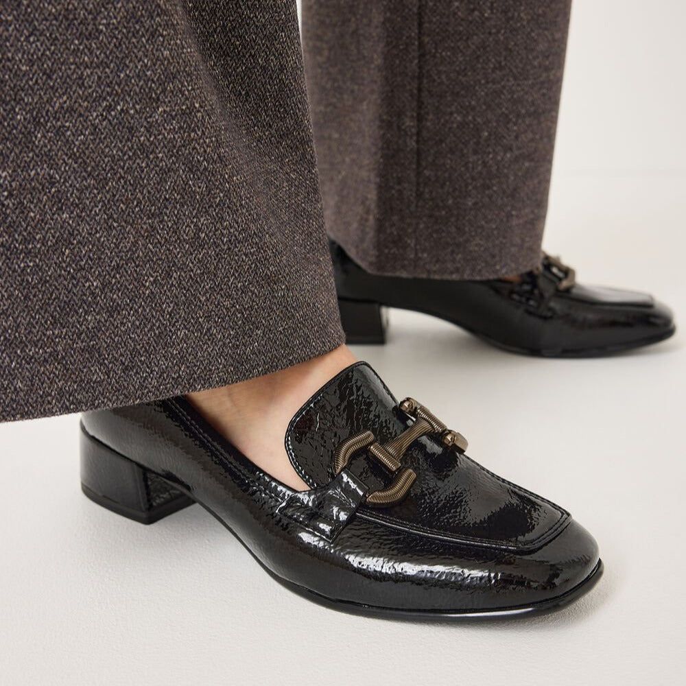 Black patent loafers worn with brown pants on a white background