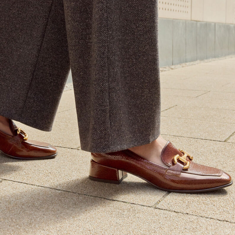 Brown loafers with gold accents worn with gray trousers on a pavement.