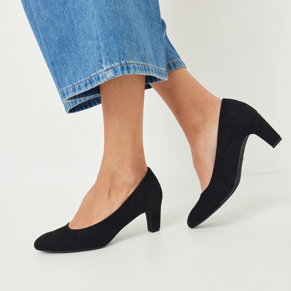 Black pointed-toe block-heel pumps worn with blue jeans, shown from the calves down on a clean white studio background.