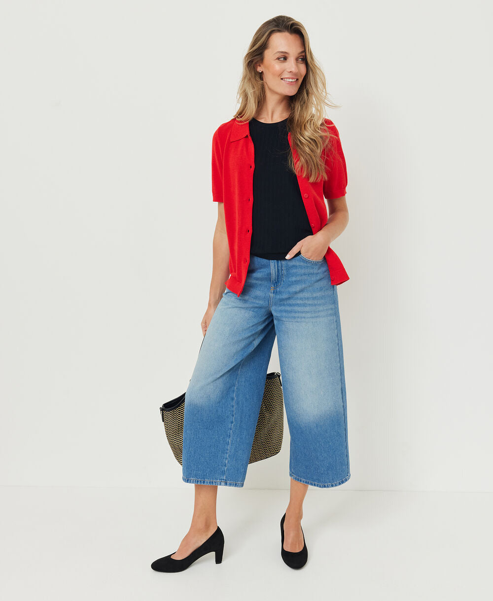 A woman modeling a vibrant red short-sleeve button-up shirt over a black top, paired with wide-leg blue denim jeans, black heels, and holding a dark olive green bag against a clean white studio background.