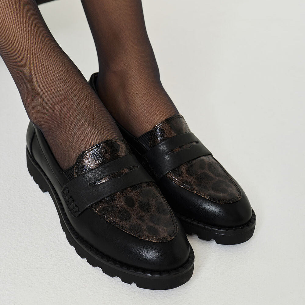 A pair of black leather penny loafers with brown and black leopard print panels, worn with sheer black tights on a light gray studio background.