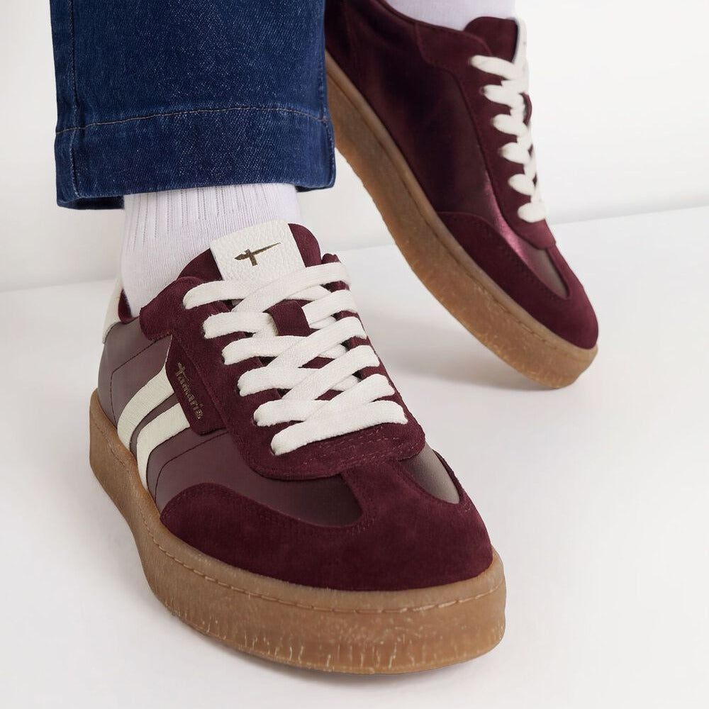 A pair of low-top platform sneakers in burgundy suede and leather with white laces, white side stripes, and a gum sole, shown on a model wearing white socks and blue jeans.