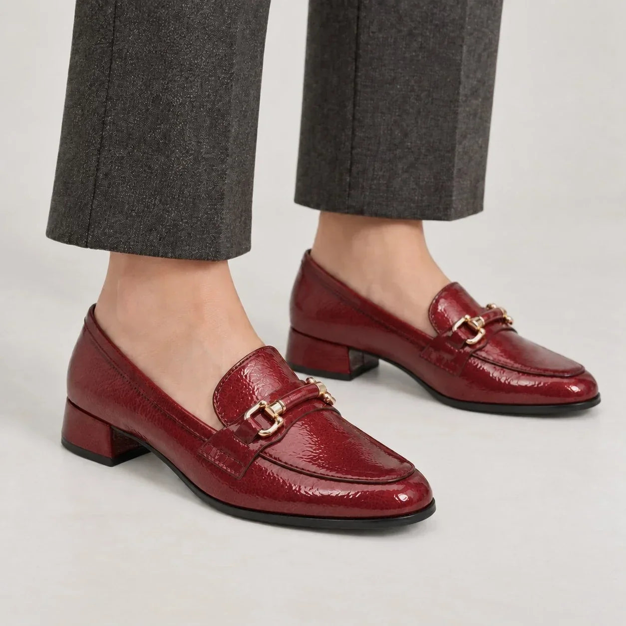 Marco Tozzi dark red patent-effect loafers with gold-tone trim, worn with grey trousers