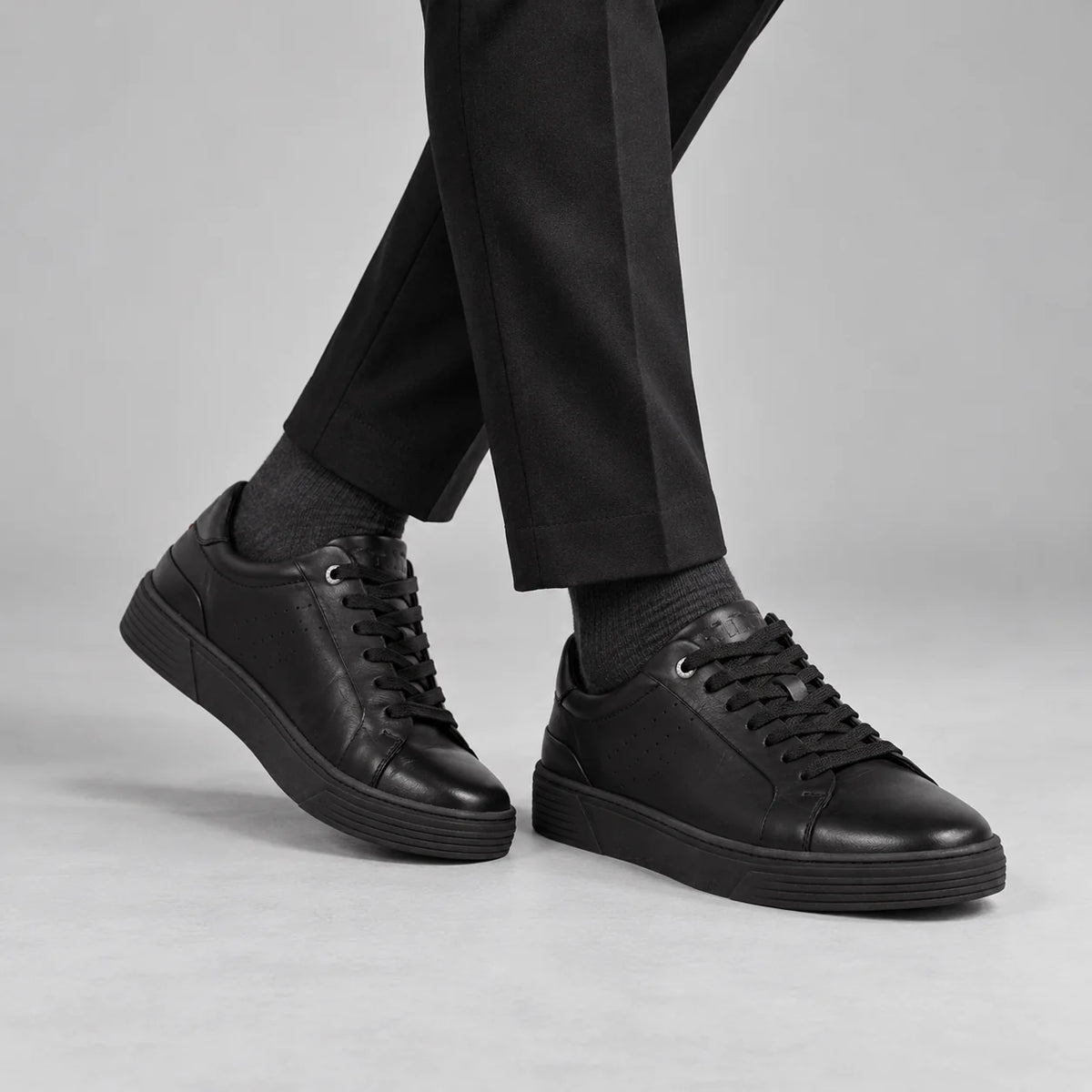 Marco Tozzi men's black leather trainers styled with black tailored trousers and charcoal socks in a neutral studio.
