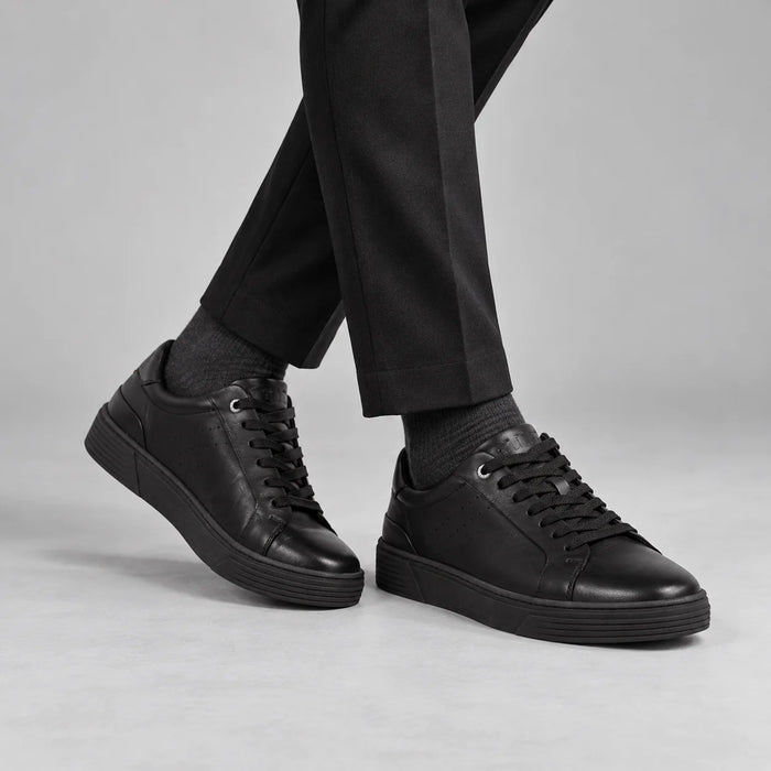 Marco Tozzi men's black leather trainers styled with black tailored trousers and charcoal socks in a neutral studio.
