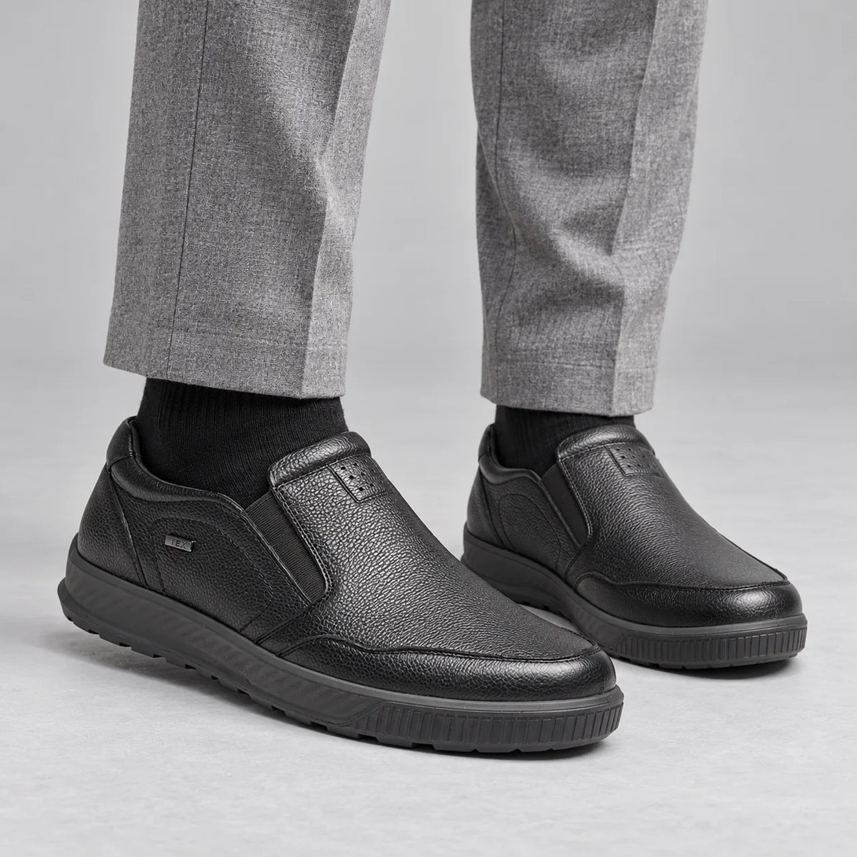 G-Comfort A-951 men's black leather slip-on shoes worn with light grey trousers and black socks in a grey studio.