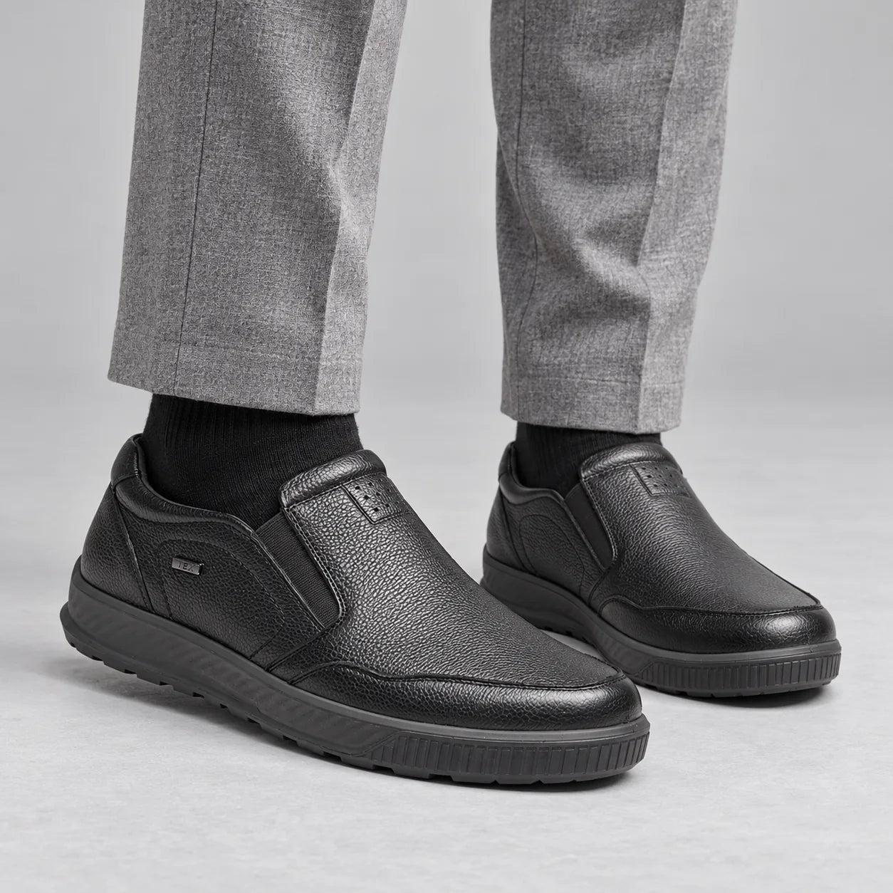 G-Comfort A-951 men's black leather slip-on shoes worn with light grey trousers and black socks in a grey studio.
