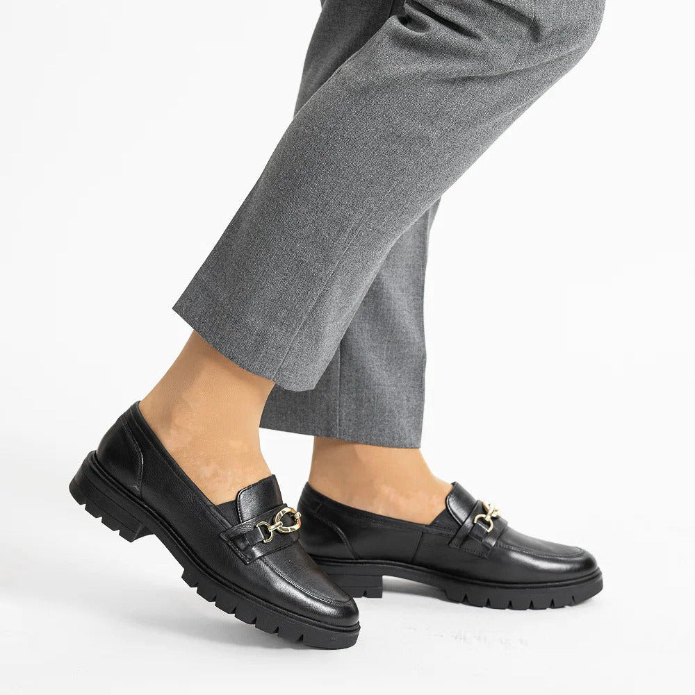 Black leather loafer-style shoes with a small gold horsebit detail, worn with gray trousers against a clean white background.