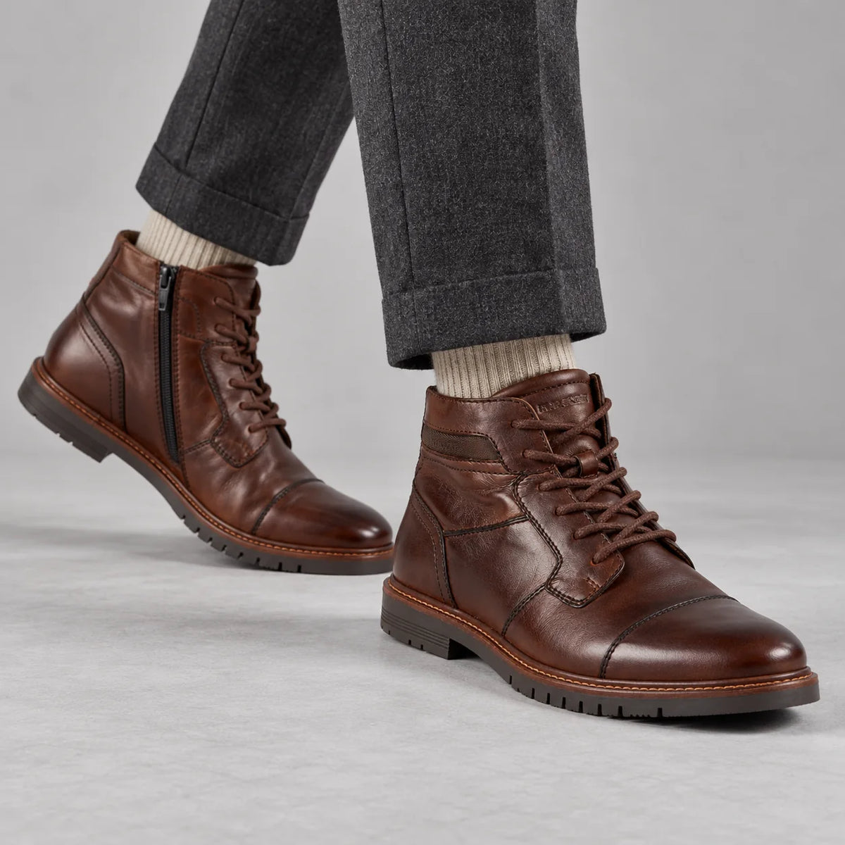 Marco Tozzi men's dark brown cap-toe boots worn with charcoal trousers and oatmeal socks in a grey studio.