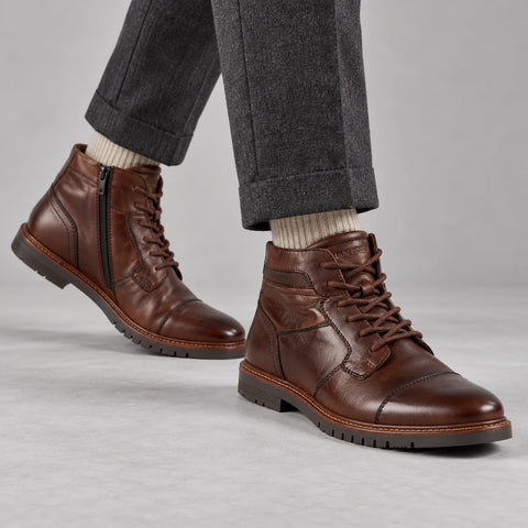Marco Tozzi men's dark brown cap-toe boots worn with charcoal trousers and oatmeal socks in a grey studio.