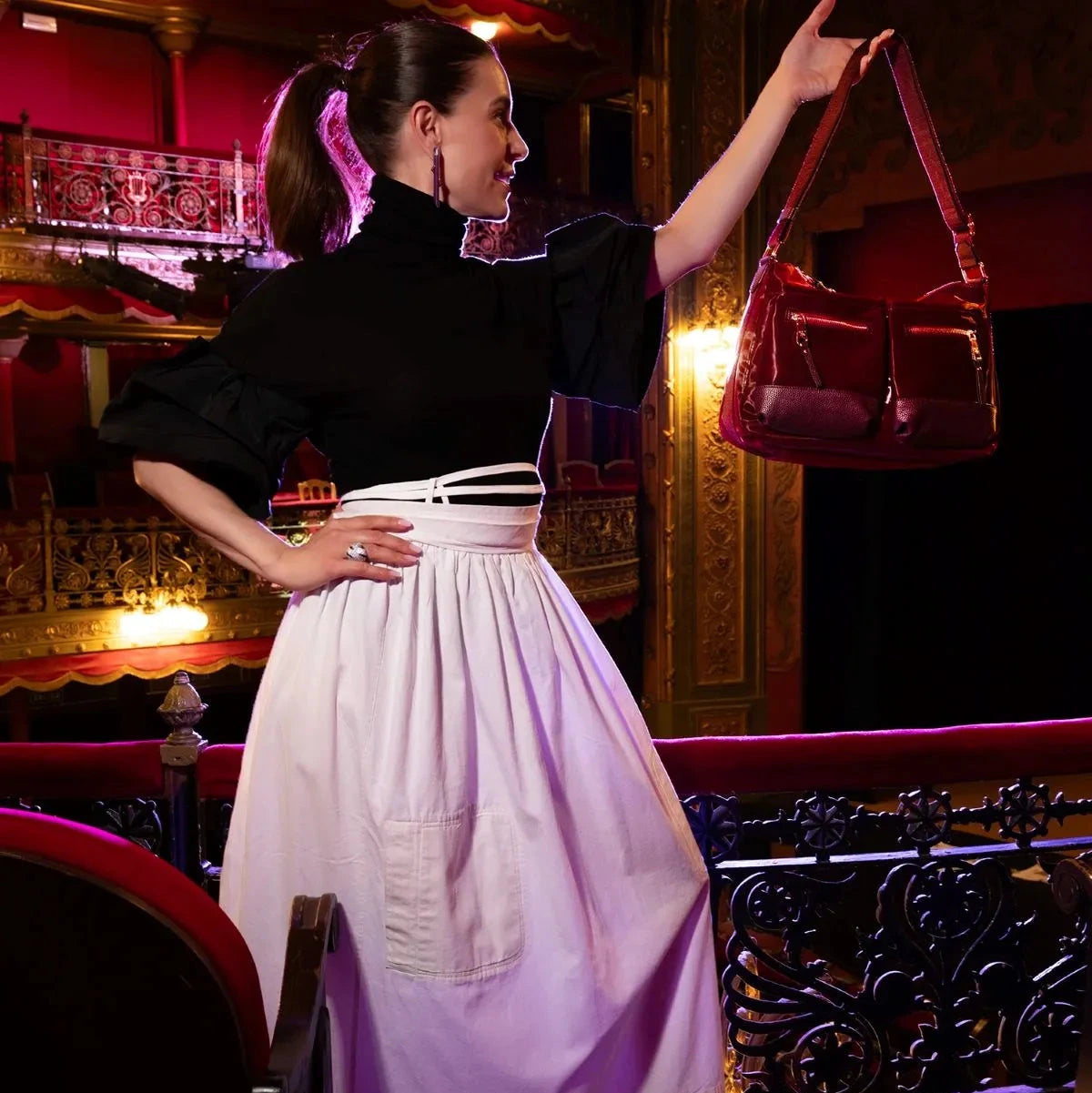 Woman in a black top and white skirt holding a red handbag in an ornate room.