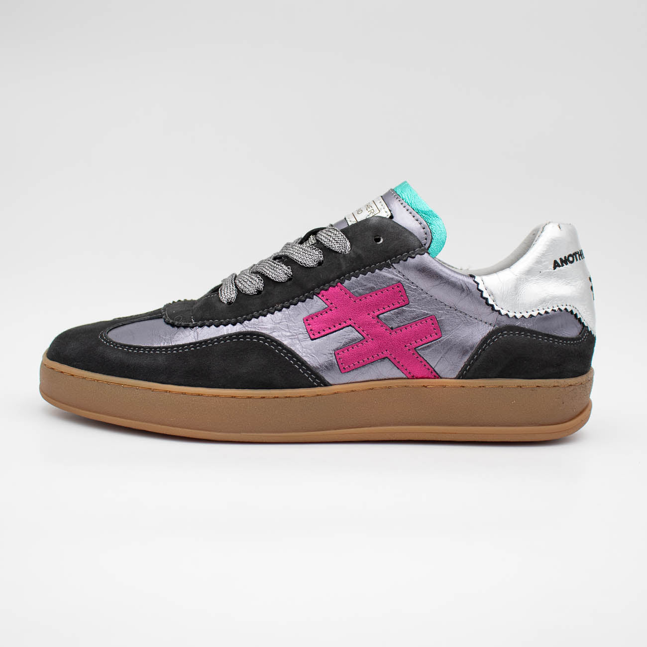 A low-top sneaker shown in profile against a light gray background. The shoe features a black suede toe cap, a metallic silver side panel with a prominent hot pink stylized 'E' logo, gray laces, a turquoise tongue accent, and a brown gum sole.
