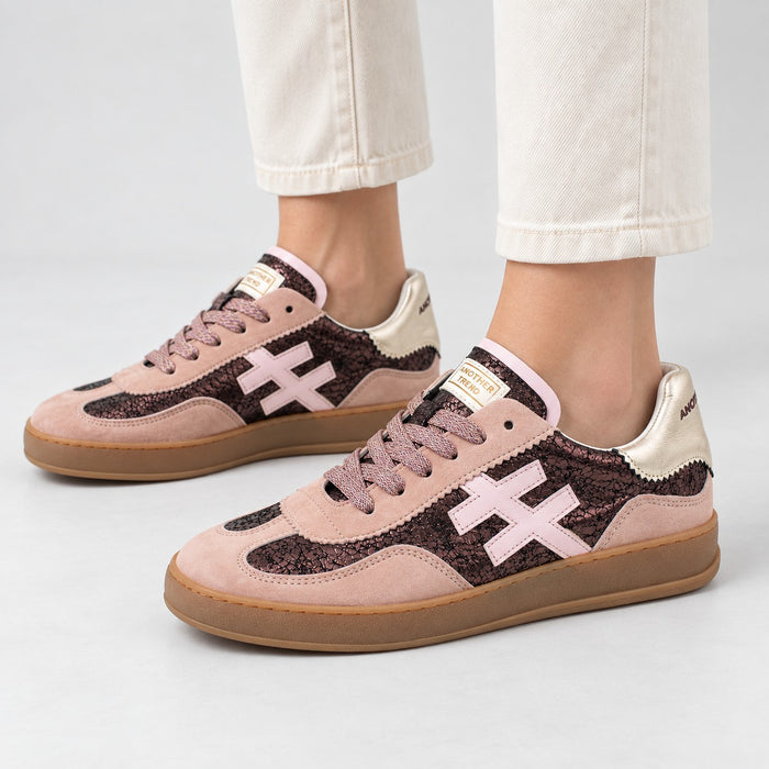 A pair of low-top sneakers worn with off-white cropped pants, shown from the side on a light gray studio background. The shoes have a dusty pink suede base with dark brown textured overlays, a light pink cross logo, and a gum brown sole.