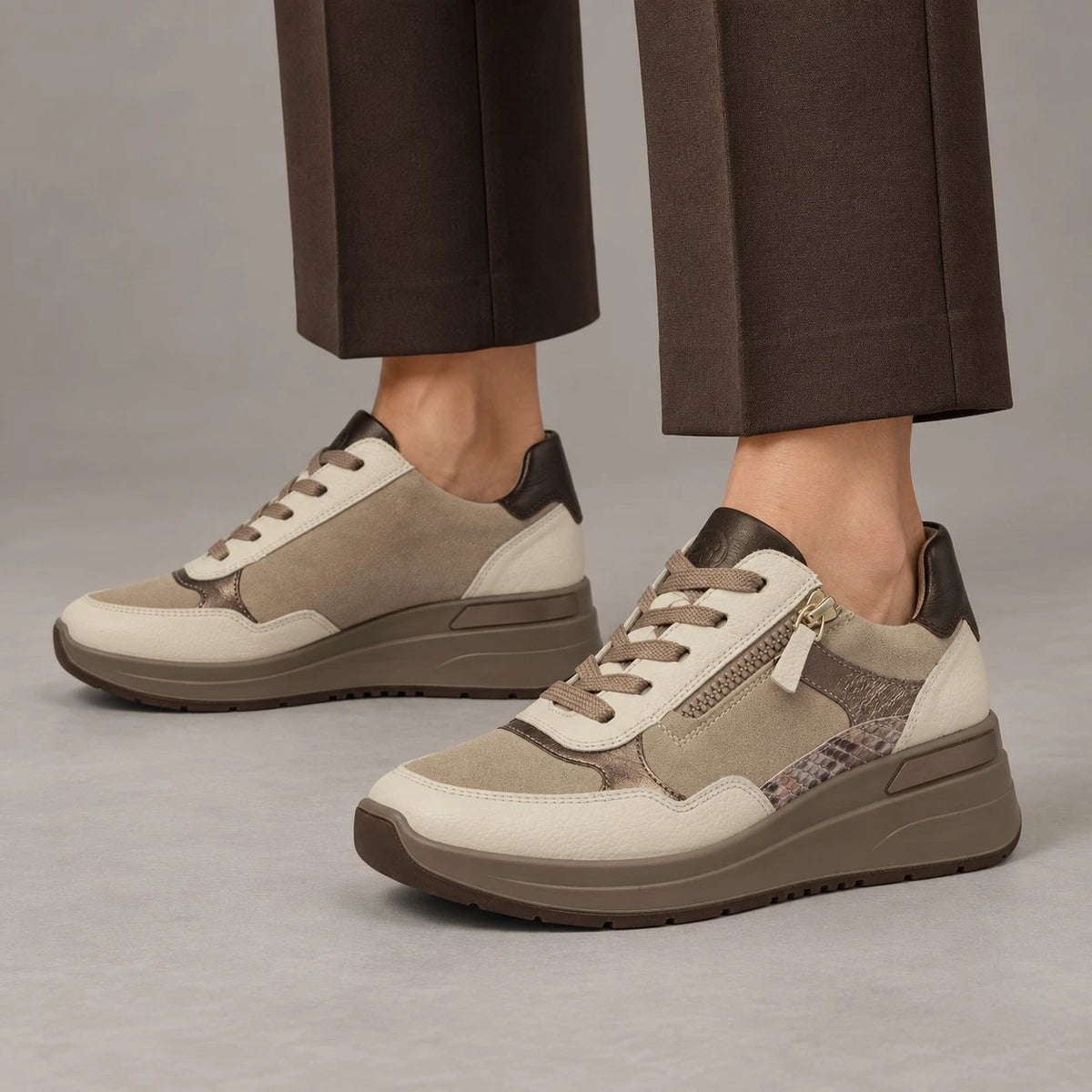 A pair of low-top platform sneakers in beige, brown, and gray tones, shown on a model wearing dark brown trousers against a neutral gray studio background.