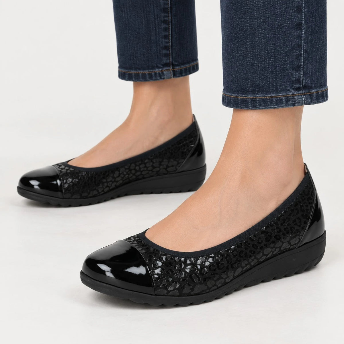 A pair of women's black slip-on flats with a glossy patent toe cap and a textured crocodile-embossed upper, shown on a model's feet wearing blue jeans against a light gray studio background.
