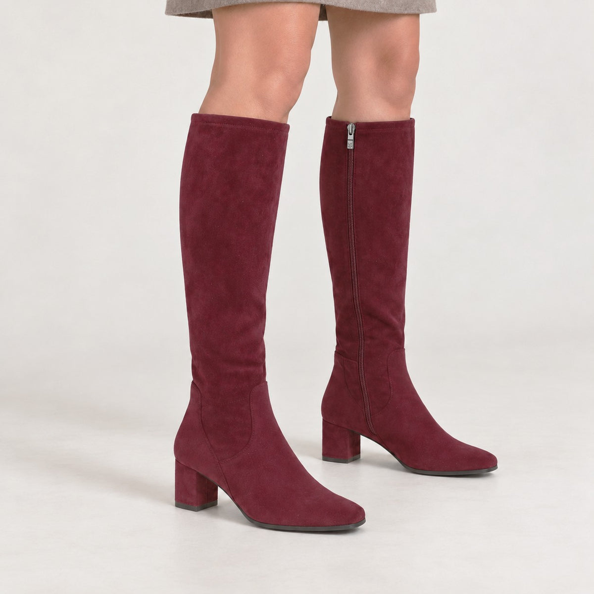 A pair of knee-high suede boots in burgundy, shown on a model against a light gray studio background.