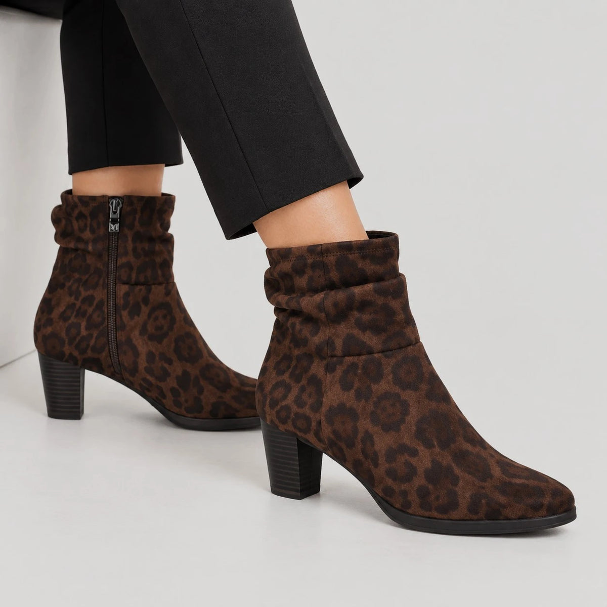 A pair of brown leopard-print ankle boots with a stacked block heel, worn with black trousers against a light gray studio background.