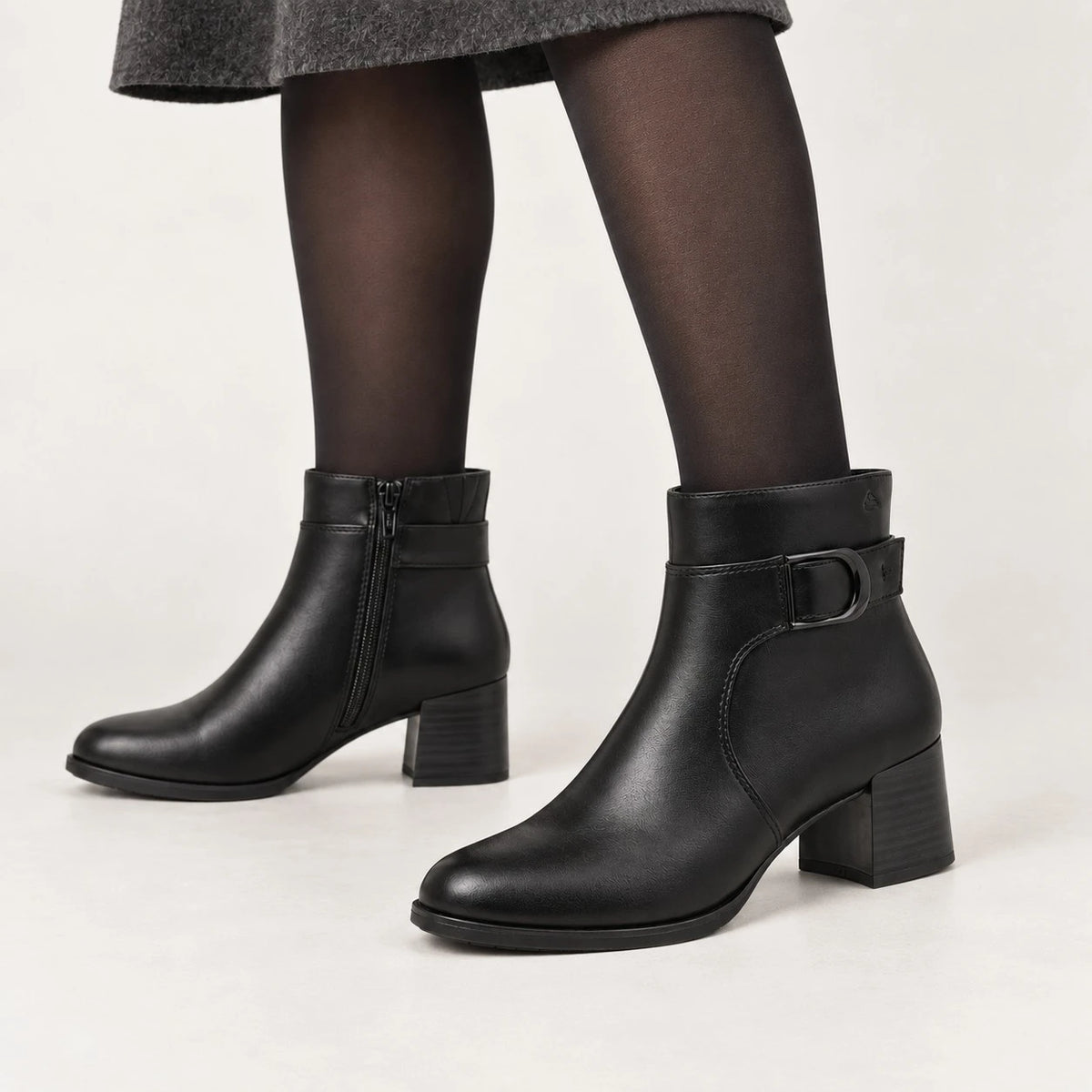 Black leather ankle boots with a block heel and side buckle detail, shown on a model wearing black tights against a light gray studio background.