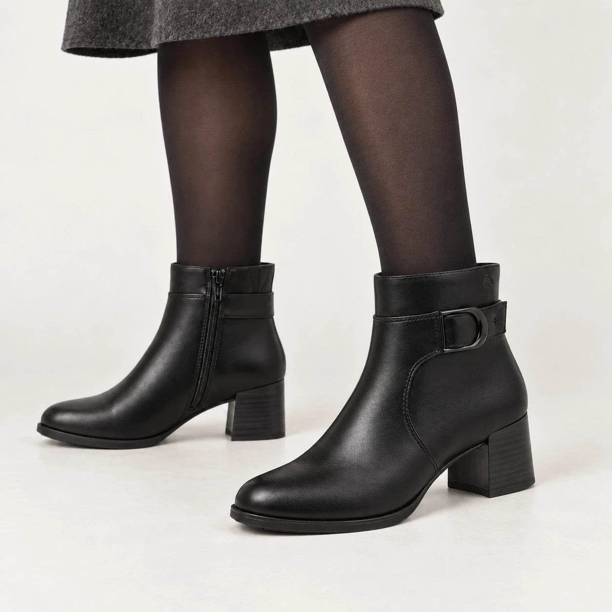 Black leather ankle boots with a block heel and side buckle detail, shown on a model wearing black tights against a light gray studio background.