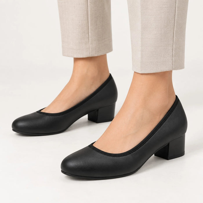 A pair of black low-heeled pumps with a rounded toe, shown on a model wearing light beige trousers against a clean white studio background.