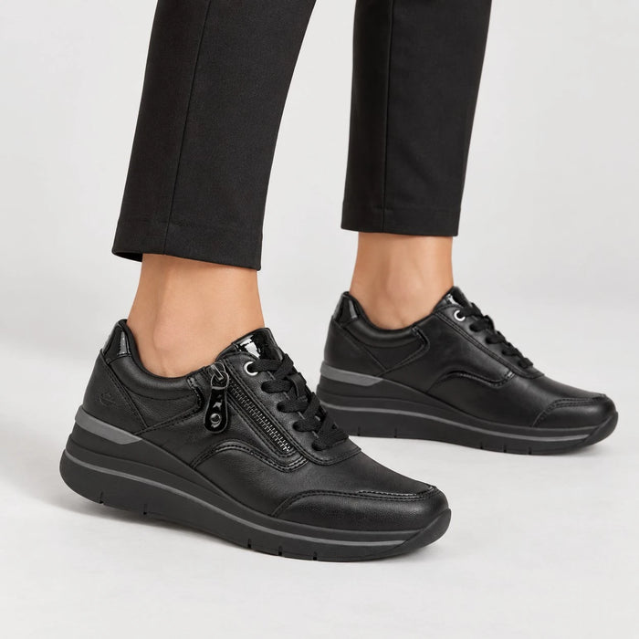 A pair of black leather low-top sneakers with a side zipper and black laces, shown on a model wearing black trousers against a light gray studio background.