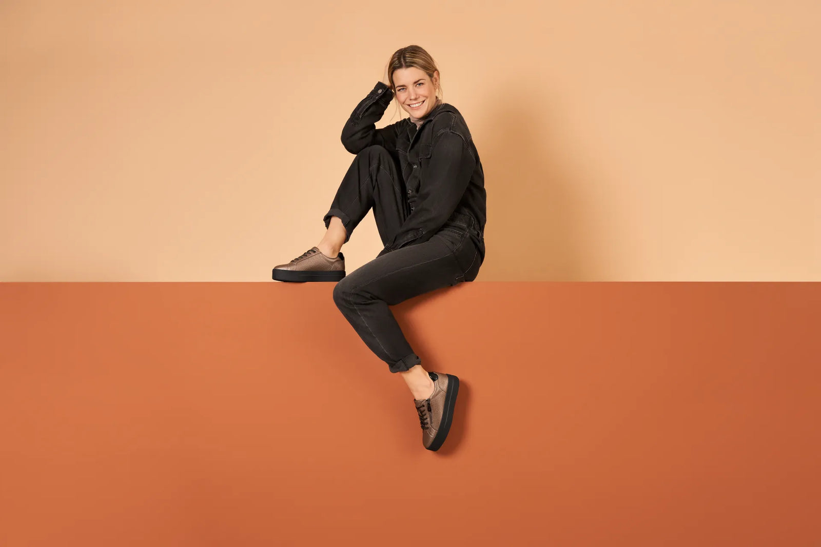 Person sitting on a brown platform wearing black clothing against a beige wall.