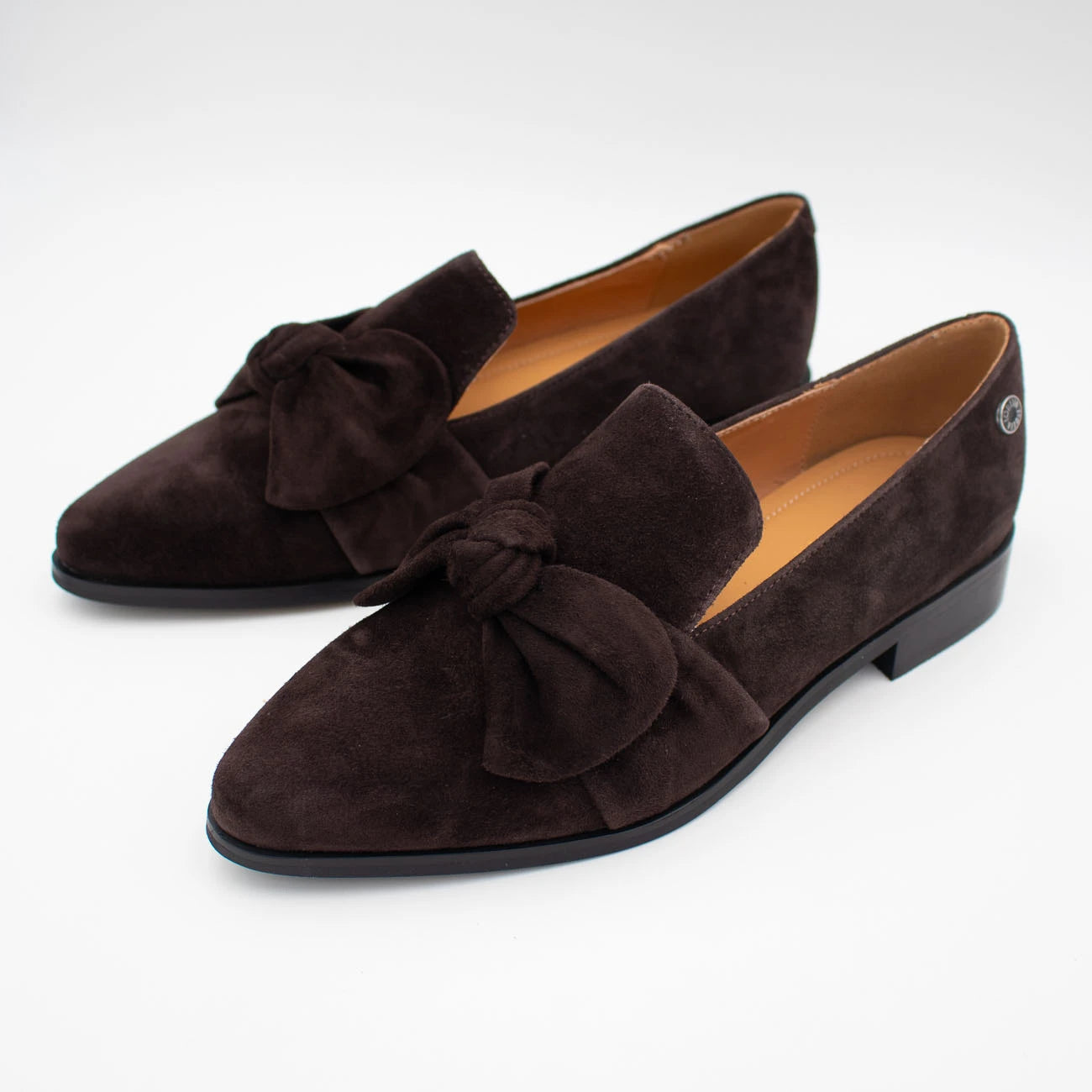A pair of dark brown suede loafers with large bow details on the toe, shown on a clean white background.