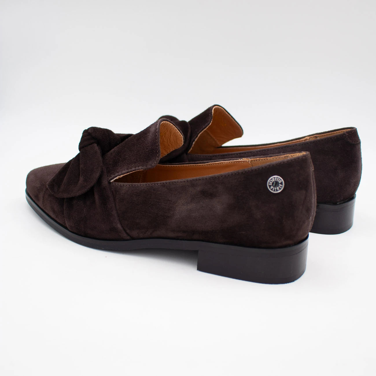 A pair of dark brown suede loafers with a knotted bow detail across the vamp, shown from a side-rear angle on a light gray background.