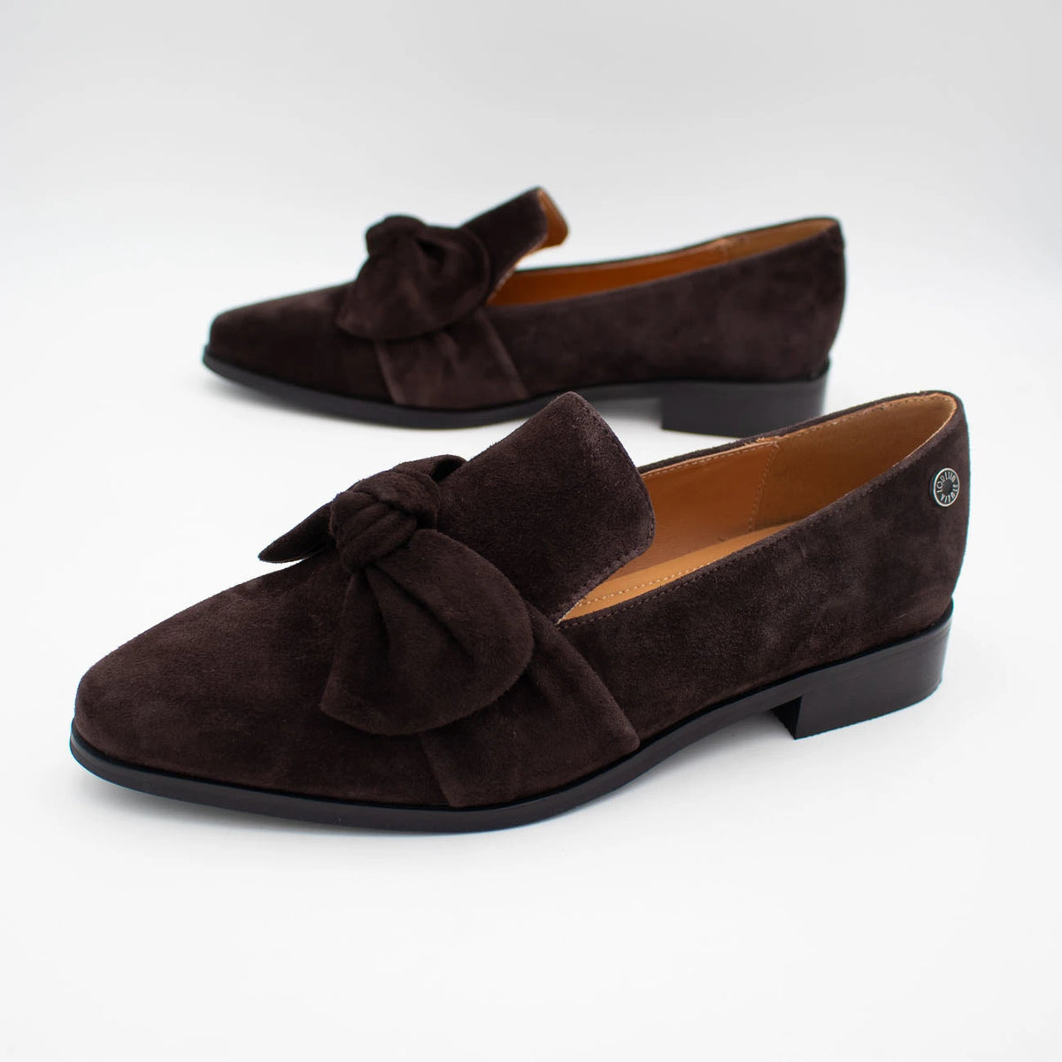 A pair of dark brown suede loafers with a bow detail on the toe, displayed on a clean white background.