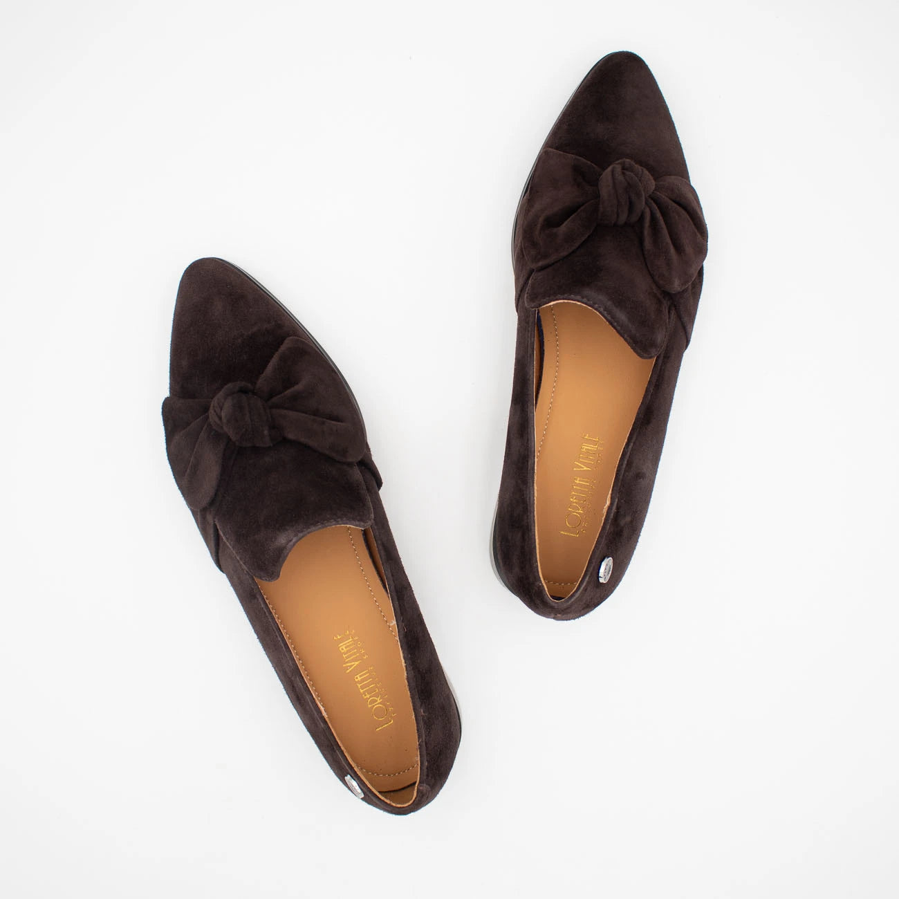A pair of dark brown suede slip-on loafers with pointed toes and decorative bows on the toes, shown from a top-down view on a white background. The brand name 'Misty May' is visible on the tan insoles.