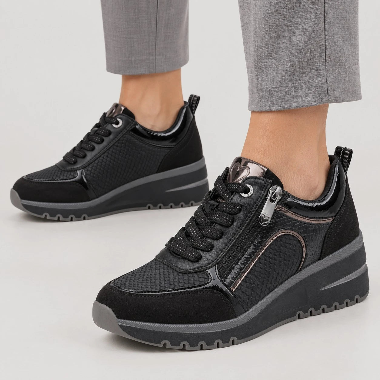 A pair of women's black platform sneakers with textured panels, side zippers, and chunky soles, shown on a model wearing gray trousers against a light gray studio background.