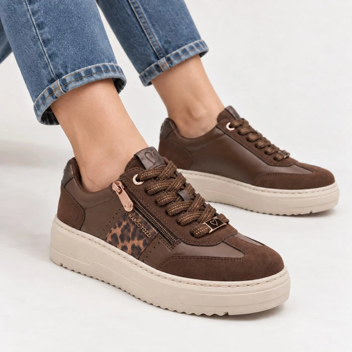 A pair of women's brown platform sneakers with a leopard print accent panel and side zipper, shown on a model wearing blue jeans against a light gray background.