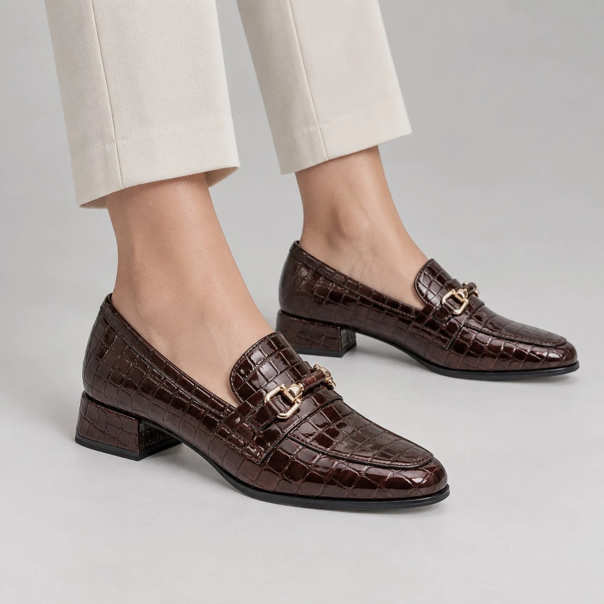 A pair of women's dark brown crocodile-embossed loafers with gold-tone buckle details, shown on a model wearing off-white trousers against a light gray studio background.