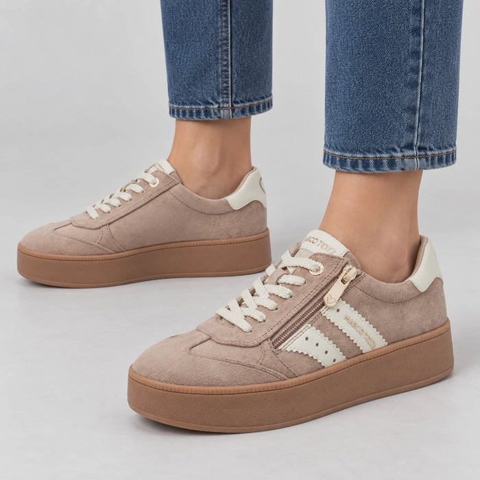 A pair of low-top platform sneakers in taupe suede with cream accents, shown on a model wearing blue jeans against a light gray studio background.