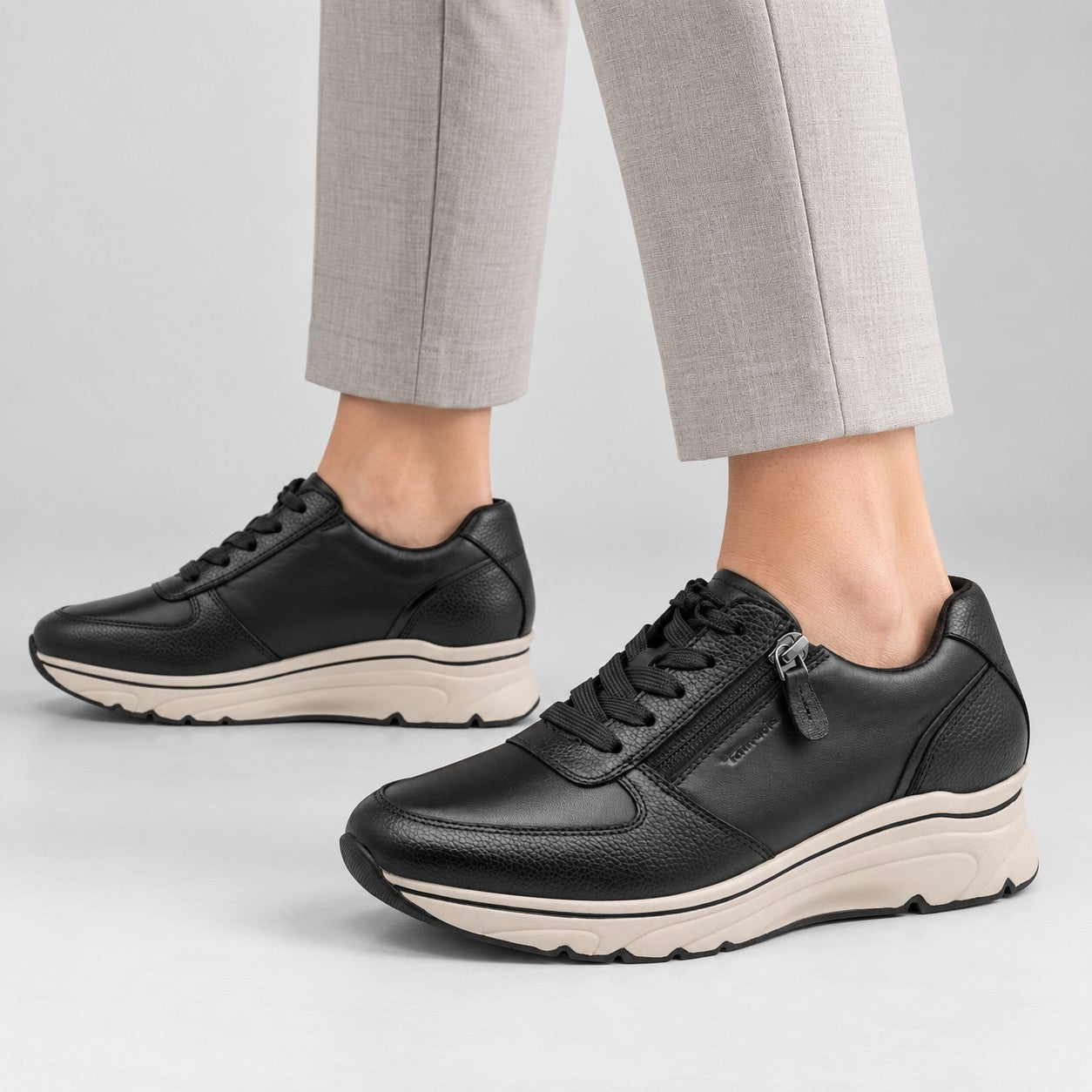 A pair of black leather low-top sneakers with side zippers, worn with light gray trousers against a light gray studio background.