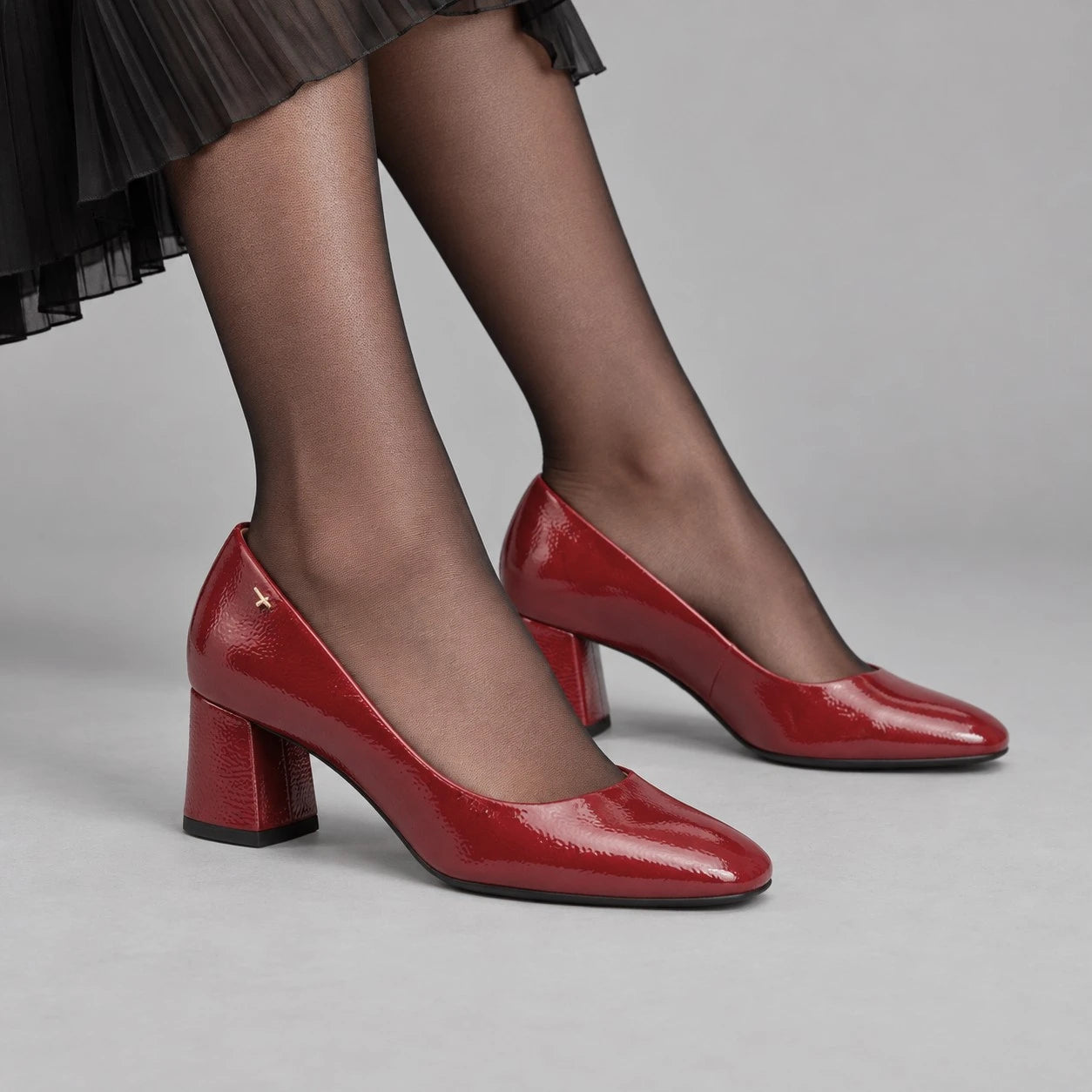 A pair of red patent leather block-heel pumps worn by a model with sheer black tights and a pleated skirt, shown against a neutral gray studio background.