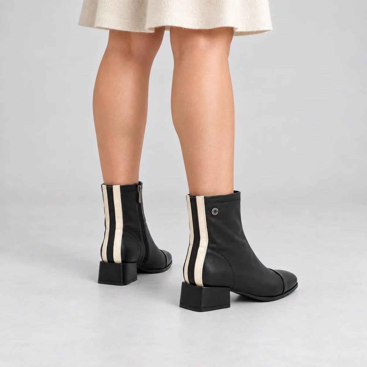 Black leather ankle boots with vertical cream stripes on the outer side, shown on a model from the back against a light gray studio background.