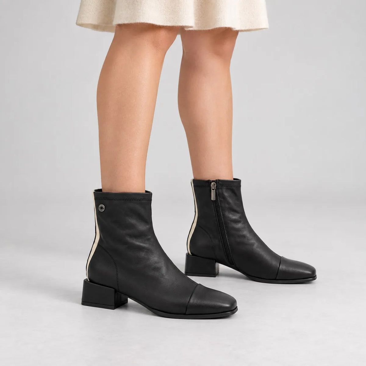 A pair of black leather ankle boots with low block heels, shown on a model wearing a light beige skirt against a light gray studio background.