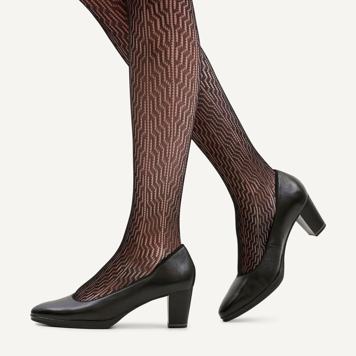 Black pointed-toe block-heel pumps worn with brown and black patterned tights, shown on a model's legs against a light off-white background.