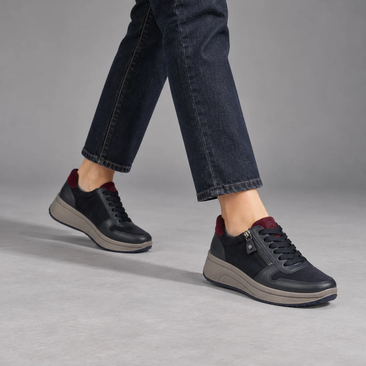 Ara Sapporo navy and merlot trainers worn with dark indigo jeans in a walking pose in a grey studio.