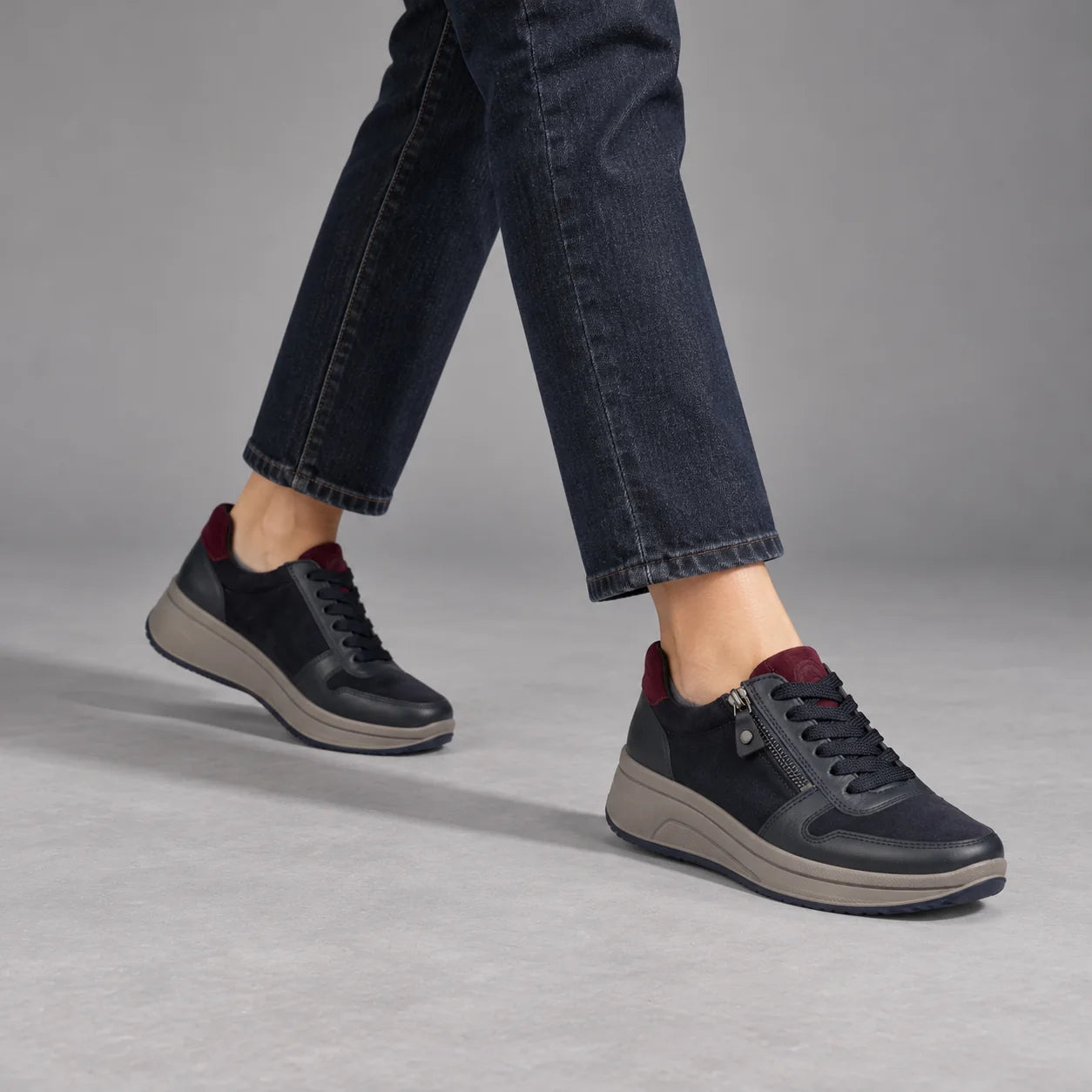 Ara Sapporo navy and merlot trainers worn with dark indigo jeans in a walking pose in a grey studio.