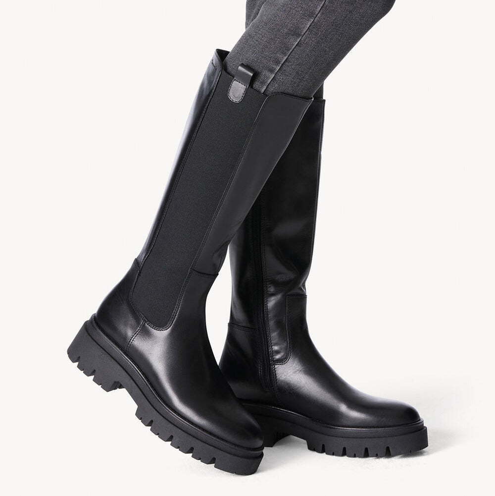Black knee-high boots worn with gray pants on a white background