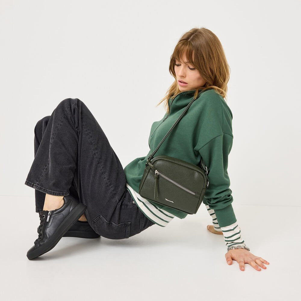 Woman sitting on the floor with a green bag, wearing a green sweater and dark pants.