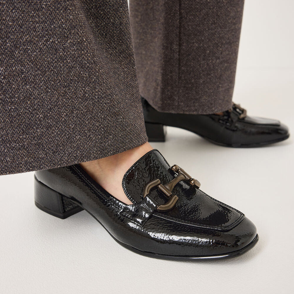 Black patent leather loafer-style loafers with a low block heel and a decorative horsebit hardware detail on the vamp, shown worn with brown textured trousers against a light gray studio background.