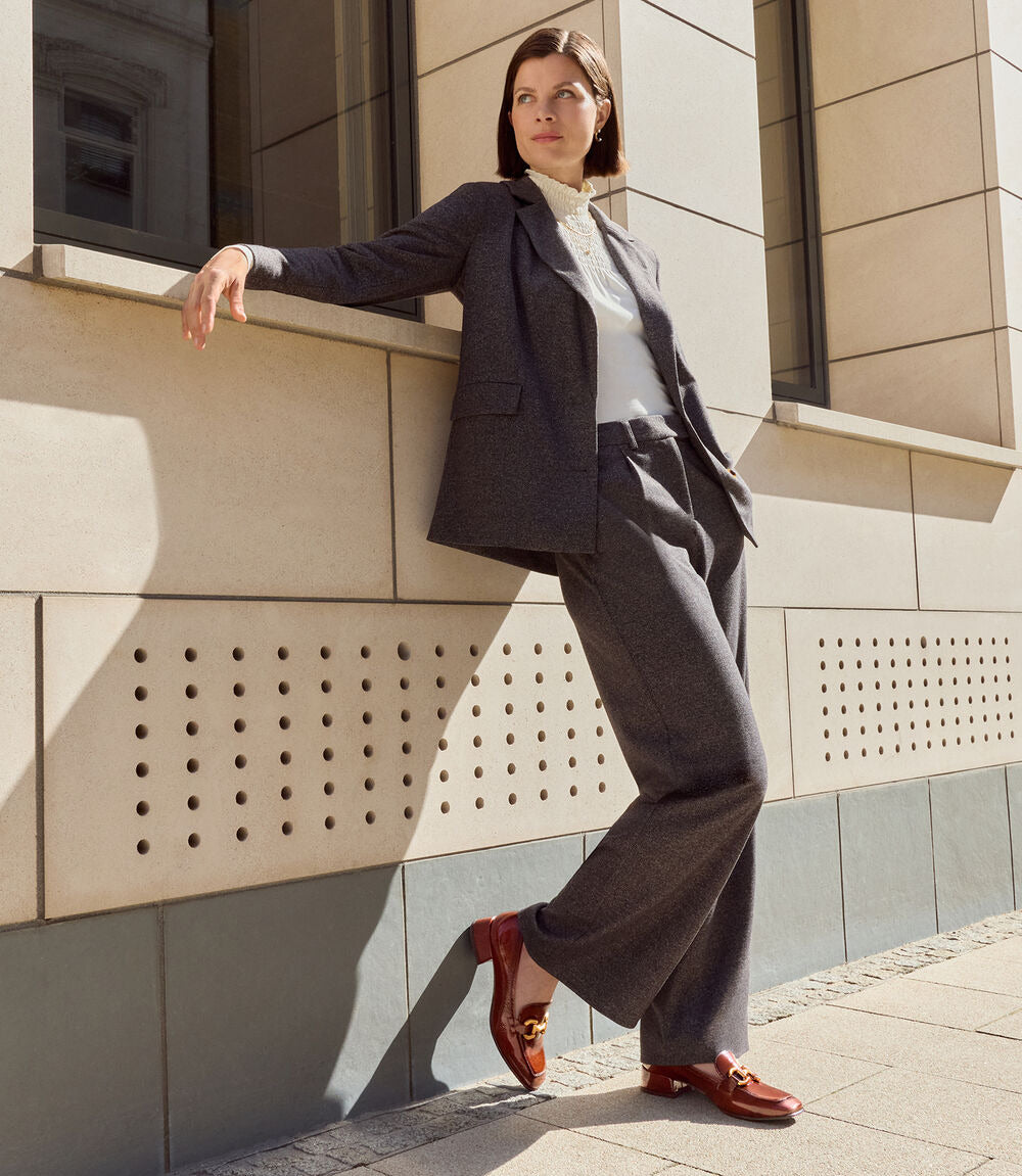 A woman models a dark gray two-piece suit consisting of a blazer and wide-leg trousers, paired with a white shirt and brown loafers, leaning against a beige stone building wall.