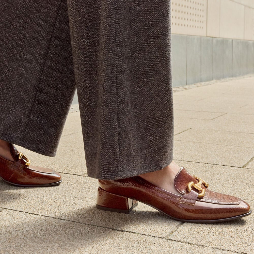 A close-up of a person wearing brown leather loafers with gold-tone horsebit detailing, paired with wide-leg taupe trousers, walking on a paved outdoor walkway.