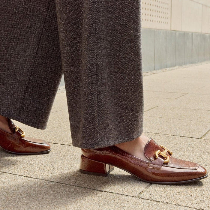 A close-up of a person wearing brown leather loafers with gold-tone horsebit detailing, paired with wide-leg taupe trousers, walking on a paved outdoor walkway.