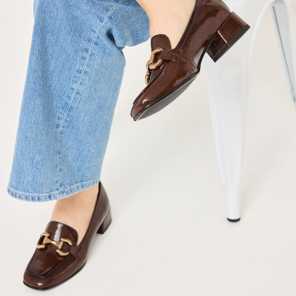 Brown loafers with gold accents worn by a person sitting on a white chair.
